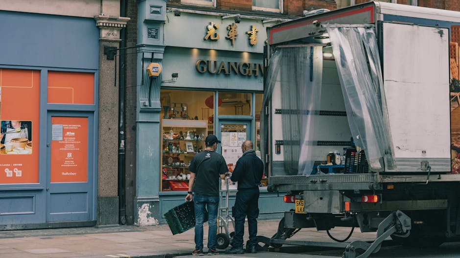 Two movers from Harringay Removals, dressed in dark clothing, are preparing to load a truck outside a shop with a sign that reads 'Guang.' One mover is lifting a black plastic crate, while the other is standing nearby, guiding the process. The truck's side panel is open, revealing a clear plastic sheet draped over the interior, and various packing supplies and equipment are visible inside. The scene takes place on a city street with a grey building façade, featuring large windows displaying products and a partially visible orange door adjacent to the shop entrance. The image captures the packing and loading stage of a home relocation or furniture transport, with the movers and their vehicle positioned on the pavement in daylight.