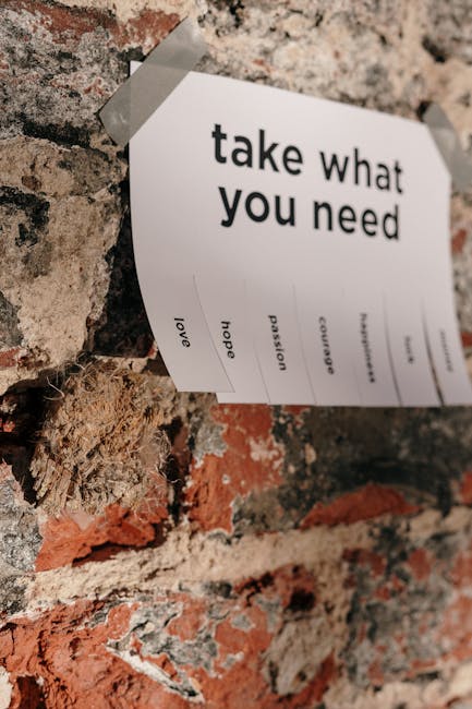 A close-up photograph of a white paper sign with printed black text that reads 'take what you need' taped onto a textured brick wall with visible rough patches and a mix of red and beige bricks. The paper appears to have several small words underneath the main message, including 'love,' 'hope,' 'passion,' and 'encourage,' suggesting a motivational or supportive message. The scene is well-lit, highlighting the contrast between the smooth paper and the uneven brick surface. This type of decor could be found in a home or community space. In the context of house removals or moving services, this image might represent a personal touch or emotional farewell element during the packing or sorting process, as offered by companies like Harringay Removals.
