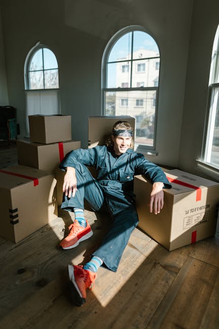 A person dressed in a blue jumpsuit, sitting on the wooden floor of a bright room with large arched and rectangular windows, surrounded by several closed cardboard boxes sealed with red and black tape, indicative of packing for a house move. The individual is wearing orange sneakers with blue socks and has a headband, appearing relaxed and casual. The room features white walls and natural daylight streaming in through the windows, which reveal a view of neighboring buildings. This scene illustrates the packing and loading process involved in a home relocation, with boxes ready for transport. The presence of boxed belongings and the interior setting aligns with services offered by companies like Harringay Removals, focused on furniture transport and packing during moving and removals.