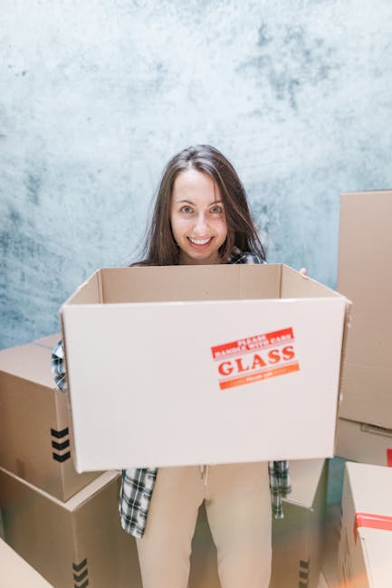A person dressed in a blue jumpsuit, sitting on the wooden floor of a bright room with large arched and rectangular windows, surrounded by several closed cardboard boxes sealed with red and black tape, indicative of packing for a house move. The individual is wearing orange sneakers with blue socks and has a headband, appearing relaxed and casual. The room features white walls and natural daylight streaming in through the windows, which reveal a view of neighboring buildings. This scene illustrates the packing and loading process involved in a home relocation, with boxes ready for transport. The presence of boxed belongings and the interior setting aligns with services offered by companies like Harringay Removals, focused on furniture transport and packing during moving and removals.