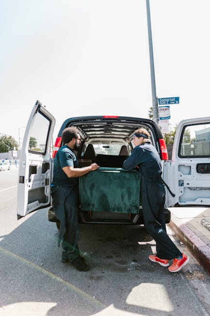 Two movers, wearing dark uniforms, are carefully loading a large, green-shaped upholstered piece of furniture into the back of a white cargo van parked on a paved street. The van’s rear doors are fully open, revealing an interior space equipped for home relocation activities, including a protective blanket draped over the furniture to avoid damage during transport. The scene is outdoors under a partly cloudy sky, with a street sign and some trees visible in the background, indicating a typical urban neighbourhood setting. The movers are using proper lifting techniques and standing on either side of the item, coordinating as part of the furniture transport process. Nearby, a sidewalk and a curb are visible, consistent with a typical house removals environment, with some administrative or packing equipment possibly out of frame. This image illustrates the careful packing and loading process involved in professional home removals, as provided by companies like Harringay Removals, supporting the broader context of packing, logistics, and furniture transport for house moves.