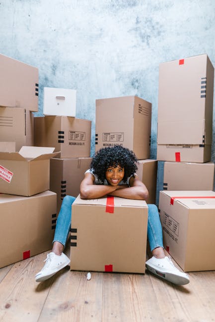 A young woman with curly black hair, wearing a white T-shirt, blue jeans, and white sneakers, is sitting on a hardwood floor surrounded by numerous cardboard boxes of various sizes, some sealed with red packing tape and others open with packing materials partially visible. She is smiling and resting her arms on a medium-sized box directly in front of her, with her legs spread out and feet flat on the floor. The scene is set inside a room with a plain, light-colored textured wall in the background. The boxes are stacked and arranged around her, indicating a packing or home relocation process, typical of furniture transport and moving preparations. This image illustrates the packing and loading aspects of house removals, showcasing the organization and handling of belongings during a home move, as often managed by companies like harringayremovals.co.uk.
