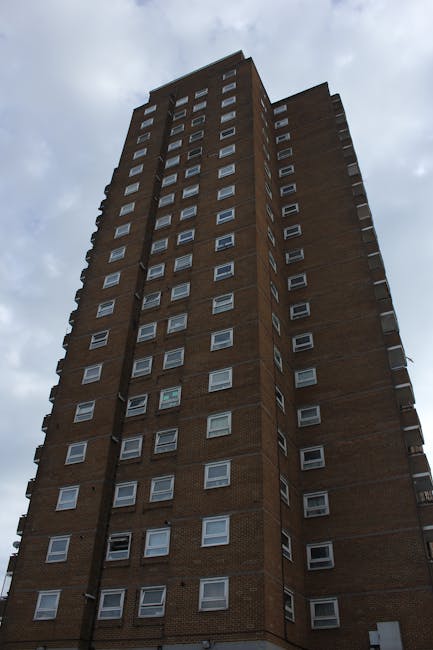 A tall multi-storey block of flats constructed from reddish-brown bricks, with multiple white-framed windows arranged in a grid pattern, extending upward into a cloudy sky. The building's exterior appears weathered and utilitarian, characteristic of urban residential properties in the UK. The perspective of the image emphasizes the height of the structure, with no foreground visible. This type of building is typical of the Harringay Ladder estate area, relevant to house removals and home relocation services offered by companies like Harringay Removals. The image captures the entire façade from ground level to the roof, showing the uniformity of window placements and the building's imposing presence in an urban environment, often requiring careful planning for access during moving or furniture transport processes.