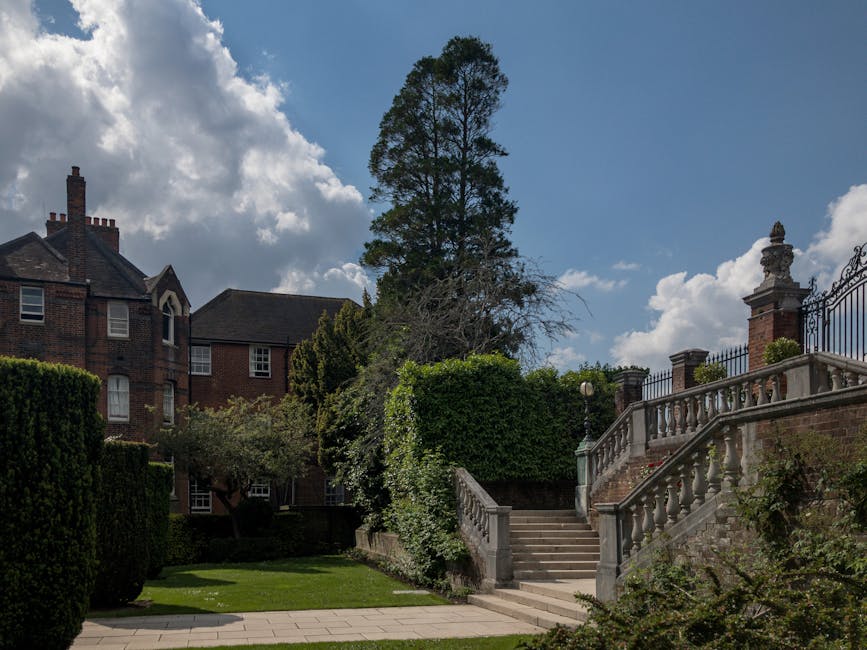 A residential property with brick buildings and classic Victorian-style architecture, featuring multiple chimneys and windows. In the foreground, there are stone steps leading up to a gated entrance with decorative stone balustrades and brick pillars topped with ornamental features. Surrounding the steps are well-maintained, neatly pruned green bushes and trees, including a tall coniferous tree extending into the bright blue sky with scattered white clouds. The surrounding garden area showcases a paved pathway and a lush, green lawn. The scene is well-lit with natural daylight, highlighting the exterior details of the property. This setting exemplifies a typical home suitable for house removals and furniture transport, with clear access points, outdoor steps, and an inviting landscape for loading and unloading during a home relocation managed by Harringay Removals.