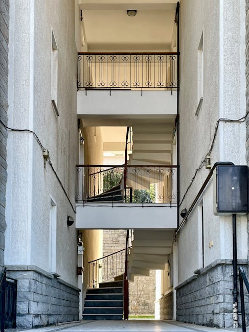 The image depicts a narrow, multi-story staircase of an apartment building with three visible floors, each featuring a small balcony with decorative black wrought iron railings. The exterior walls are light-colored, with several small rectangular windows on each level. A concrete staircase with white steps and a dark red rail connects the floors, and the ground floor shows a door at the base of the stairs. On the right side of the image, an electrical box is mounted on the wall, with electrical wiring running along the surface. The lighting appears natural, indicating daytime, with sunlight illuminating the scene. Inside the stairwell, household items such as cardboard boxes and plastic wrapping are visible, suggesting ongoing packing or disassembly related to a home removal or relocation. The setting is typical of an urban residential property, and the image aligns with house removal procedures involving navigating tight, multi-storey staircases during furniture transport and packing and moving activities, as managed by companies like Harringay Removals.