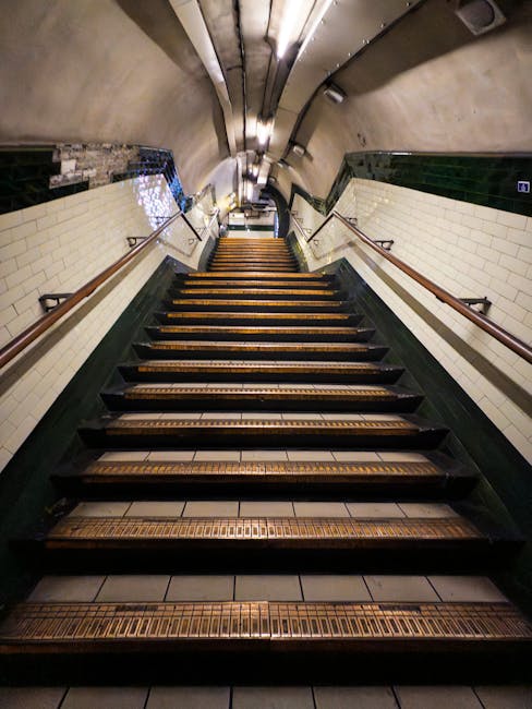 A downward view of a narrow staircase inside a building, featuring wooden steps with non-slip strips and metal railings on both sides. The staircase is enclosed by white tiled walls with dark green trim at the bottom, and the ceiling has a curved, metallic finish with lighting fixtures illuminating the area. The photograph captures the staircase during a home relocation process, with no furniture or boxes visible in the image. This setting of a confined staircase highlights common problems faced during furniture transport and moving logistics, especially when navigating tight or narrow staircases, as often handled by professional removals services like Harringay Removals.