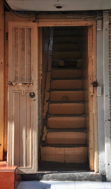 The image depicts a narrow, multi-story staircase of an apartment building with three visible floors, each featuring a small balcony with decorative black wrought iron railings. The exterior walls are light-colored, with several small rectangular windows on each level. A concrete staircase with white steps and a dark red rail connects the floors, and the ground floor shows a door at the base of the stairs. On the right side of the image, an electrical box is mounted on the wall, with electrical wiring running along the surface. The lighting appears natural, indicating daytime, with sunlight illuminating the scene. Inside the stairwell, household items such as cardboard boxes and plastic wrapping are visible, suggesting ongoing packing or disassembly related to a home removal or relocation. The setting is typical of an urban residential property, and the image aligns with house removal procedures involving navigating tight, multi-storey staircases during furniture transport and packing and moving activities, as managed by companies like Harringay Removals.