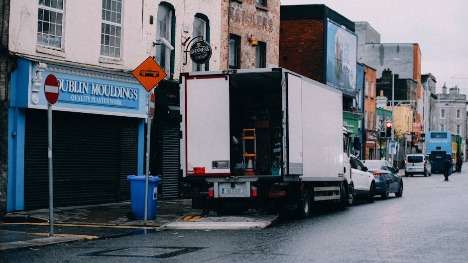 A white box truck with its rear doors open is parked on a city street outside a building with a blue sign reading 'Dublin Mouldings' and a Guinness logo, indicating a premises related to plaster work. The truck is positioned partly on the sidewalk and pavement, with a trolley inside visible for loading or unloading. Nearby, there is a blue waste bin and concrete barriers on the pavement. The street is lined with a variety of small business storefronts painted in different colours; some shops have signs, and there is a pedestrian crossing with a red traffic light visible in the distance. Several other vehicles, including cars and a bus, are parked along the street or moving through the area. The environment suggests an active urban setting suitable for home removals, furniture transport, and packing and moving activities, with a focus on efficient loading processes facilitated by the professional services of [COMPANY_NAME]. The lighting appears natural, indicating daytime, with overcast weather contributing to diffuse light conditions.