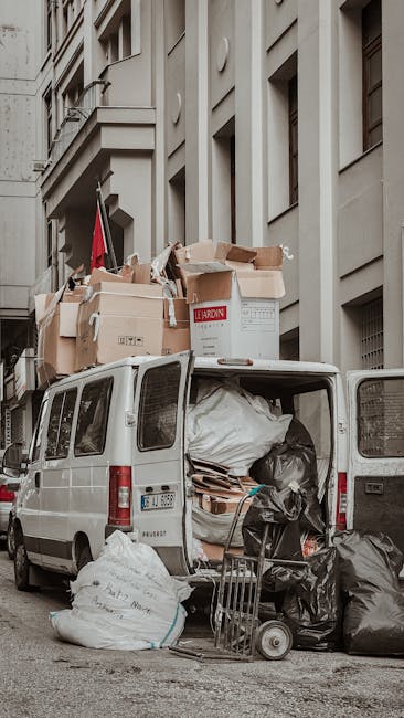 A white commercial van parked on a city street next to a multi-storey building, with its rear doors open revealing a large collection of packed boxes, black and white plastic garbage bags, and various packing materials inside. Several cardboard boxes, some open and some sealed, are stacked on the roof of the van, with additional boxes and packaging supplies loaded onto the vehicle. A small foldable trolley and a few black trash bags are on the pavement beside the van, indicating an ongoing loading process typical of home relocation or furniture transport. The building features beige or light-colored stone façade with multiple rectangular windows, some partially visible above the van. The scene captures a moment of house move logistics, with clear signs of packing and loading for a removal service, as part of a same day emergency move in Harringay, managed by Harringay Removals.