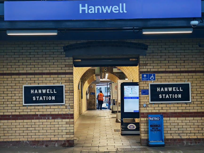 Image of the exterior entrance to Hanwell Station, displaying brickwork with two black and white signs reading 'HANWELL STATION' mounted on either side of a doorway. Above the entrance, a blue sign with white text indicates the station's name. Inside the entrance, a person wearing an orange high-visibility vest is visible near the ticket barriers, suggesting the station's active passenger area. A digital information display is positioned on the right side of the entrance, and a small blue accessibility sign is mounted nearby, indicating facilities for disabled travelers. In front of the station entrance, there are objects related to packing and moving logistics, including a blue Metro trolley and a standing sign promoting walking topics. The overall scene reflects a typical urban railway station environment, with brick architecture, signage, and associated objects used during a home relocation or furniture transport process, as coordinated through professional removals like those offered by companies such as harringayremovals.co.uk.