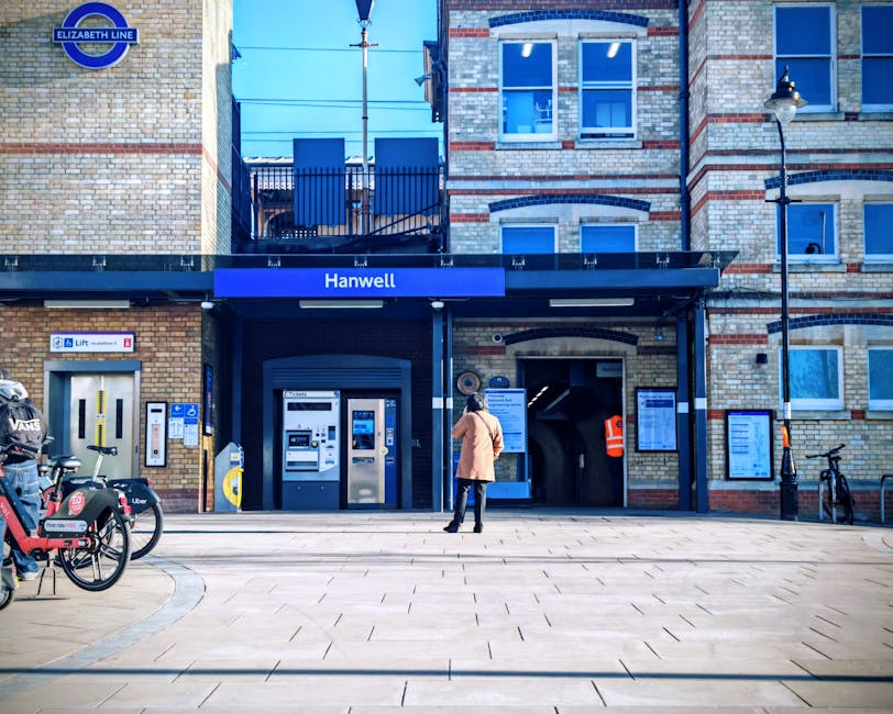 Image of the exterior entrance to Hanwell Station, displaying brickwork with two black and white signs reading 'HANWELL STATION' mounted on either side of a doorway. Above the entrance, a blue sign with white text indicates the station's name. Inside the entrance, a person wearing an orange high-visibility vest is visible near the ticket barriers, suggesting the station's active passenger area. A digital information display is positioned on the right side of the entrance, and a small blue accessibility sign is mounted nearby, indicating facilities for disabled travelers. In front of the station entrance, there are objects related to packing and moving logistics, including a blue Metro trolley and a standing sign promoting walking topics. The overall scene reflects a typical urban railway station environment, with brick architecture, signage, and associated objects used during a home relocation or furniture transport process, as coordinated through professional removals like those offered by companies such as harringayremovals.co.uk.
